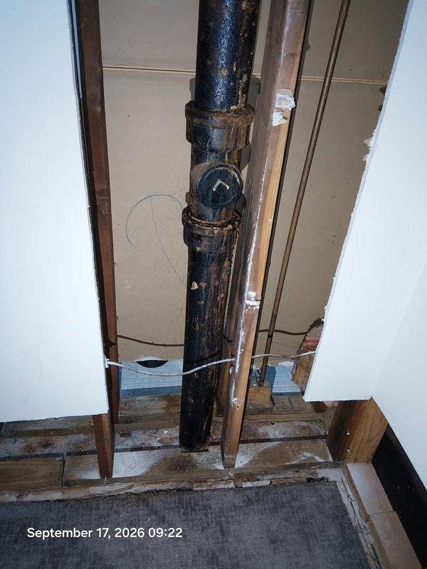 Responded to bathroom leak complaint and performed diagnostic inspection. Opened interior wall to expose plumbing infrastructure and identified deteriorated vertical waste pipe with significant corrosion and surface degradation at multiple coupling joints. Black drain pipe exhibited rust, discoloration, and aging throughout its vertical run, with visible moisture staining on surrounding floor surfaces indicating active or recent water infiltration. Documented condition of compromised pipe system, exposed wall cavity revealing wooden framing and drywall backing, and adjacent copper supply line. Inspection confirmed structural pipe failure requiring replacement to prevent continued water damage. Responded to bathroom leak complaint and performed diagnostic inspection. Opened interior wall to expose plumbing infrastructure and identified deteriorated vertical waste pipe with significant corrosion and surface degradation at multiple coupling joints. Black drain pipe exhibited rust, discoloration, and aging throughout its vertical run, with visible moisture staining on surrounding floor surfaces indicating active or recent water infiltration. Documented condition of compromised pipe system, exposed wall cavity revealing wooden framing and drywall backing, and adjacent copper supply line. Inspection confirmed structural pipe failure requiring replacement to prevent continued water damage.