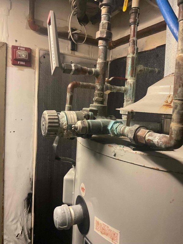 Conducted assessment for water heater replacement at Cross Ultra Lounge, documenting corroded T-valve with significant mineral buildup and oxidation on connection points. Inspection revealed an 80-gallon unit with deteriorated components and evaluated existing mechanical room configuration with copper piping showing verdigris for proper replacement planning.