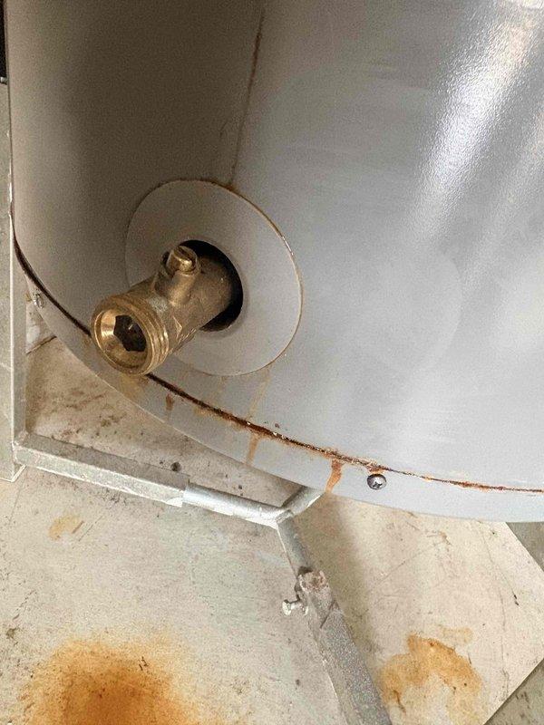 Inspected Rheem water heater previously replaced under warranty that is now exhibiting significant leakage from the bottom connection point. Documented extensive rust and corrosion around brass water connection valve, with visible water damage extending to the surrounding floor area. Determined unit requires replacement as the tank integrity has been compromised.