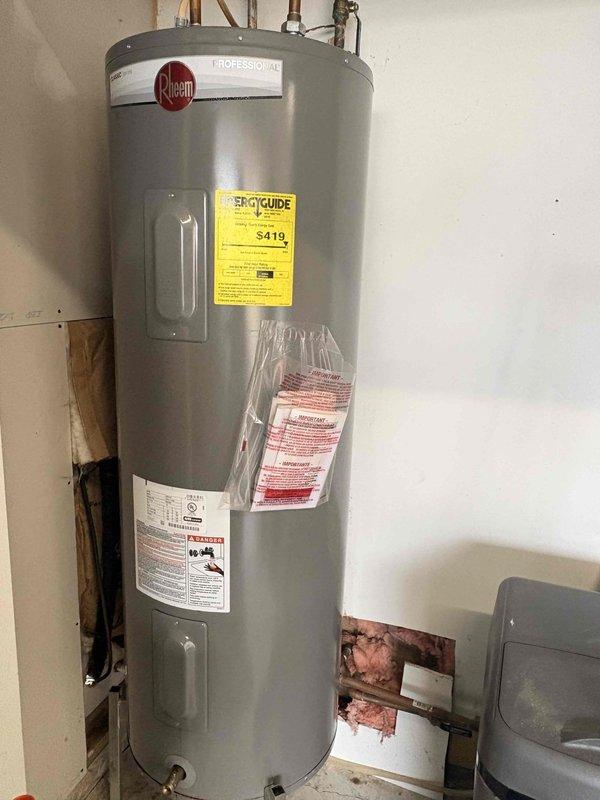 Inspected Rheem water heater previously replaced under warranty that is now exhibiting significant leakage from the bottom connection point. Documented extensive rust and corrosion around brass water connection valve, with visible water damage extending to the surrounding floor area. Determined unit requires replacement as the tank integrity has been compromised.