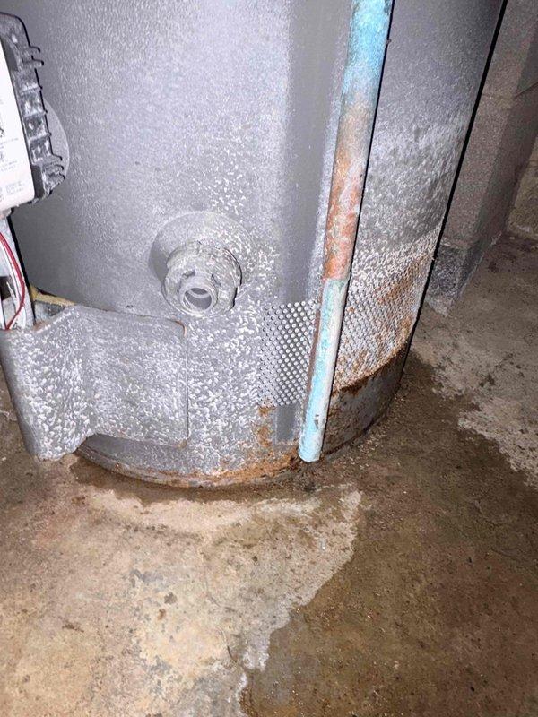 Conducted inspection of Rheem Performance water heater showing significant deterioration. Unit exhibited substantial rust formation at base where metal contacts concrete floor, with visible moisture staining indicating possible leakage. Observed copper pipes connected to top of unit, proper installation against concrete block wall, and functioning connections, though aging system shows signs of extended moisture exposure requiring attention.