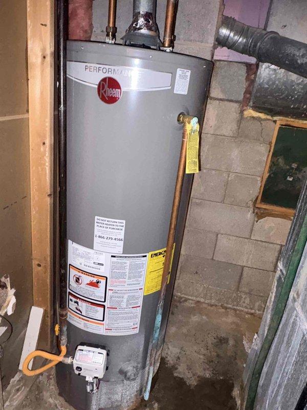 Conducted inspection of Rheem Performance water heater showing significant deterioration. Unit exhibited substantial rust formation at base where metal contacts concrete floor, with visible moisture staining indicating possible leakage. Observed copper pipes connected to top of unit, proper installation against concrete block wall, and functioning connections, though aging system shows signs of extended moisture exposure requiring attention.