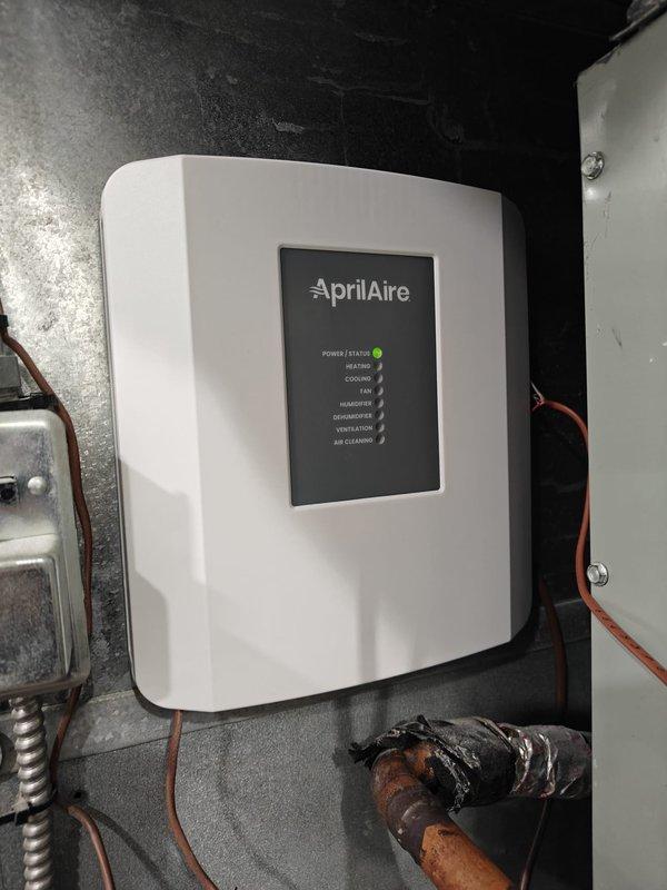 Successfully installed customer-supplied AprilAire thermostat control unit, properly connecting all required wiring (red, yellow, green, blue, white, and orange) to appropriate terminals. Verified system functionality with power indicator showing green status light illumination. Control unit now properly configured to manage heating, cooling, fan, humidity, dehumidification, ventilation, and air cleaning functions.