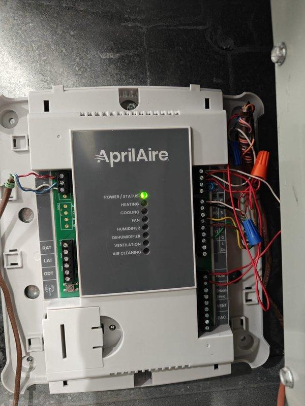Successfully installed customer-supplied AprilAire thermostat control unit, properly connecting all required wiring (red, yellow, green, blue, white, and orange) to appropriate terminals. Verified system functionality with power indicator showing green status light illumination. Control unit now properly configured to manage heating, cooling, fan, humidity, dehumidification, ventilation, and air cleaning functions.