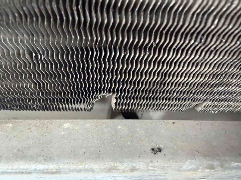 Responded to customer report of heavy water leakage from AC ducts in back office area. Upon inspection, examined evaporator coil assembly and drain pan system. Photos documented the condition of the heat exchanger fins and drain pan, which showed visible signs of moisture accumulation, dirt buildup, and mineral deposits on the horizontal drain surface beneath the corrugated aluminum fins. The drain pan exhibited discoloration and debris consistent with condensate overflow or blocked drainage. Assessed the evaporator coil fins for proper airflow and checked drain line functionality to address the reported water leakage issue.