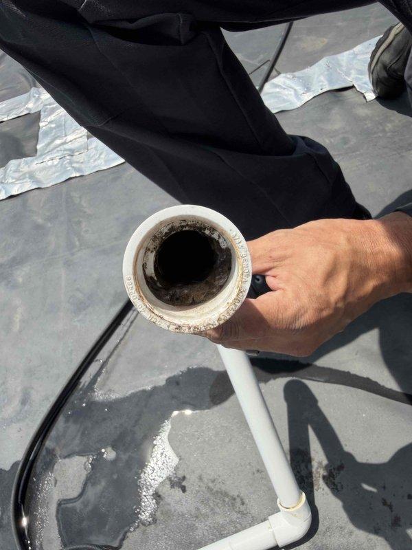 Responded to service call for reported water leak from HVAC unit. Upon inspection, found condensate drain line showing signs of blockage with visible dark staining and debris buildup inside the PVC pipe. Examined roof penetration at unit location and noted deteriorated sealing around drain line pass-through with significant rust corrosion present on equipment panel base, indicating prolonged moisture exposure. Documented existing conditions with photos showing compromised drain system and weathered equipment enclosure consistent with unit age over 10 years.