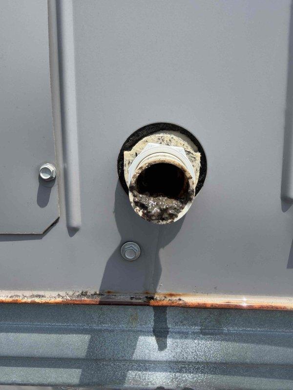 Responded to service call for reported water leak from HVAC unit. Upon inspection, found condensate drain line showing signs of blockage with visible dark staining and debris buildup inside the PVC pipe. Examined roof penetration at unit location and noted deteriorated sealing around drain line pass-through with significant rust corrosion present on equipment panel base, indicating prolonged moisture exposure. Documented existing conditions with photos showing compromised drain system and weathered equipment enclosure consistent with unit age over 10 years.