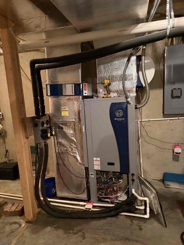 Completed installation of a new 5-ton Water Furnace 5-series geothermal heat pump system in basement mechanical room. System includes main heat pump unit mounted on concrete block wall with insulated refrigerant lines, supply and return ductwork with reflective insulation, electrical disconnect panel, and integrated controls. All ductwork connections sealed with metallic tape, PVC condensate drainage installed, and system startup procedures performed with temperature verification readings confirming proper operation at 530.3°F on heat mode testing.