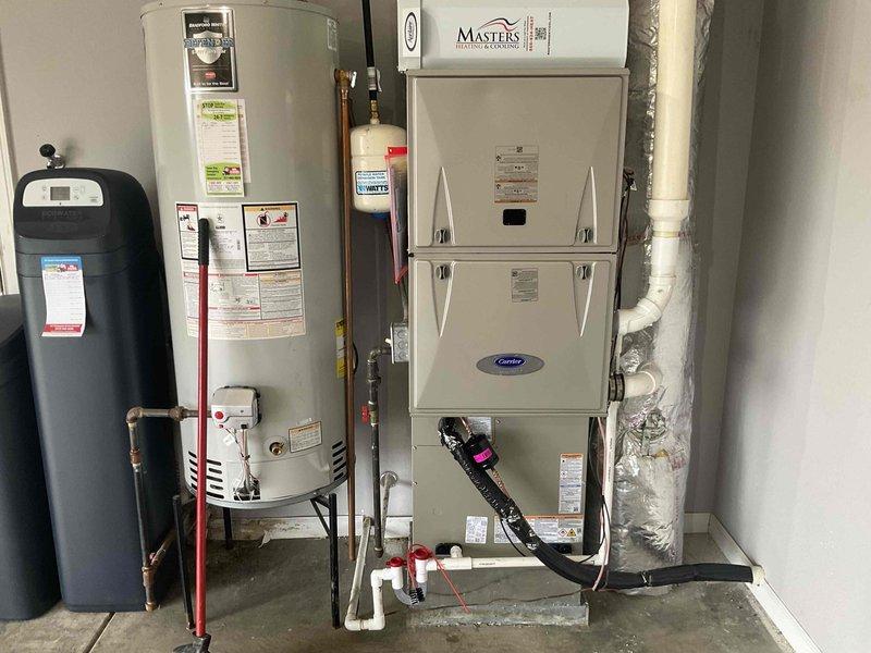 Completed full HVAC system installation including Carrier brand furnace unit installed in utility room with proper gas line connections and ductwork integration, outdoor condenser unit positioned on gravel pad against exterior brick wall with electrical connections, and water heater with expansion tank and copper piping properly integrated into the system. All equipment secured, leveled, and tested for proper operation with refrigerant lines connected between indoor and outdoor components.