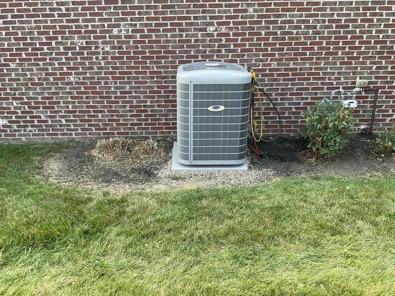 Completed full HVAC system installation including Carrier brand furnace unit installed in utility room with proper gas line connections and ductwork integration, outdoor condenser unit positioned on gravel pad against exterior brick wall with electrical connections, and water heater with expansion tank and copper piping properly integrated into the system. All equipment secured, leveled, and tested for proper operation with refrigerant lines connected between indoor and outdoor components.