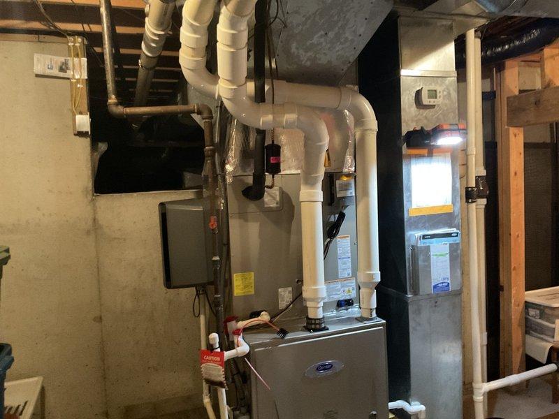Completed full HVAC system installation in residential basement mechanical room. Installed new Carrier furnace unit with proper venting using white PVC piping routed vertically through ceiling joists, copper refrigerant lines, and electrical connections to existing panel. Configured supply and return ductwork with galvanized steel plenum connections to distribute conditioned air throughout residence. All components tested and verified operational upon completion.