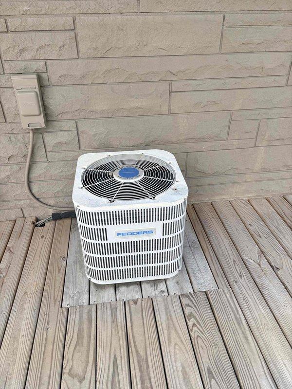 Completed seasonal preventative maintenance on two residential cooling systems. Inspected and serviced first unit, a Fedders cylindrical condensing unit located on exterior deck, verifying proper operation of fan assembly, checking electrical connections at wall-mounted outlet, and confirming adequate airflow through vertical slat venting. Performed maintenance on second unit, a hexagonal condenser positioned on ground-level pad against stone exterior wall, inspecting refrigerant lines, electrical conduit, fan grille assembly, and ensuring proper drainage around unit base. Both systems operational and running within manufacturer specifications.