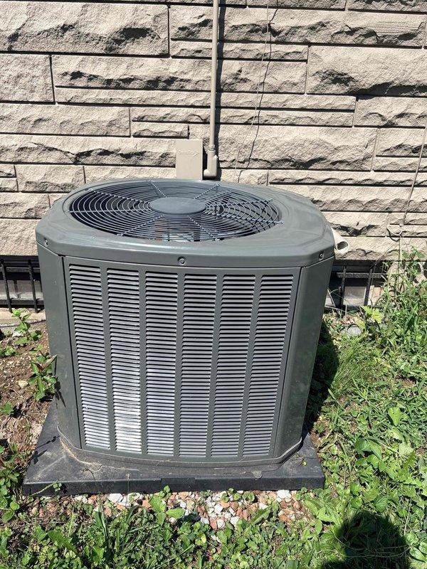 Completed seasonal preventative maintenance on two residential cooling systems. Inspected and serviced first unit, a Fedders cylindrical condensing unit located on exterior deck, verifying proper operation of fan assembly, checking electrical connections at wall-mounted outlet, and confirming adequate airflow through vertical slat venting. Performed maintenance on second unit, a hexagonal condenser positioned on ground-level pad against stone exterior wall, inspecting refrigerant lines, electrical conduit, fan grille assembly, and ensuring proper drainage around unit base. Both systems operational and running within manufacturer specifications.