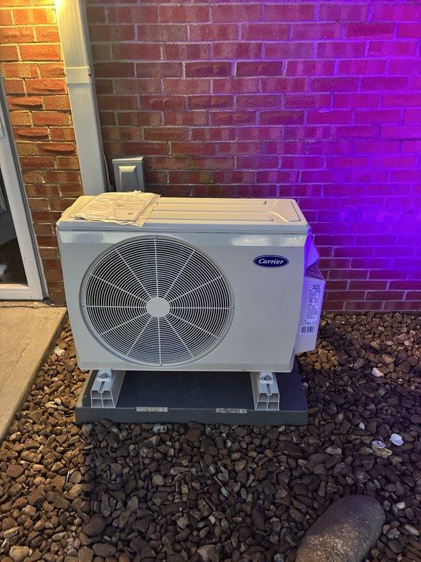 Completed installation of Carrier multizone ductless heat pump system consisting of one 37MGRAQ18CA3 Preferred Series inverter-driven outdoor condensing unit paired with two Performance Series floor console indoor units (45MBFAQ09XA3 and 45MBFAQ12XA3). Outdoor condenser was mounted on composite platform with proper elevation and positioned against exterior brick wall with surrounding decorative rock base for drainage and stability. Indoor floor console units were installed and mounted to interior walls with refrigerant line sets properly sized, routed, and connected between outdoor and indoor components. System was evacuated, pressure tested for leaks, and charged with refrigerant per manufacturer specifications. All electrical connections completed including dedicated circuits for outdoor unit and individual indoor zones. Condensate drain lines installed and tested for proper drainage. System startup performed with verification of proper operation in both heating and cooling modes acr