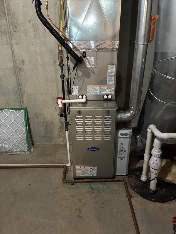 Completed full HVAC system installation in basement utility room consisting of Carrier Performance Series equipment. Installed Carrier 26SPA630W003 single-stage air conditioner (16 SEER), Carrier 58TP0B070V17--16 two-stage non-condensing gas furnace (70,000 BTU), and Carrier CVAVA3617XMA evaporator coil. Positioned furnace against concrete block wall with proper clearances and mounted metallic plenum assembly above unit. Connected refrigerant lines using copper tubing with proper brazing techniques and insulation. Installed black flexible gas line to furnace with appropriate fittings and safety shutoff valve. Ran electrical wiring and established proper 24V control circuits between outdoor condensing unit and indoor air handler. Fabricated and installed galvanized steel supply ductwork with secure connections and proper sealing. Installed white PVC condensate drain line with appropriate pitch and trap configuration. Mounted return air filter rack with green frame housing mesh filter me