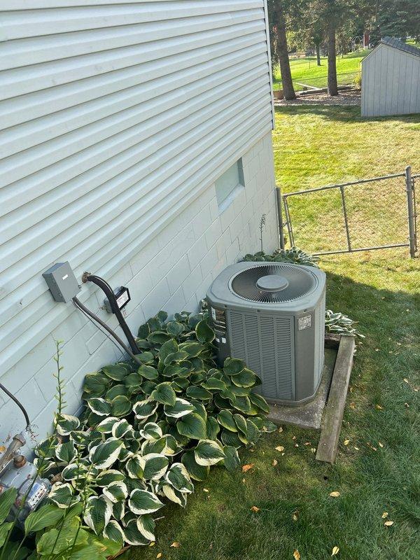 Assessed 23-year-old A/C condenser unit positioned on deteriorating wooden platform that has partially collapsed on one corner. Unit is mounted on a hillside against concrete wall with significant rot in the support structure. Recommended complete system replacement rather than attempting to level existing platform, as moving the aged unit for repairs would not be cost-effective.