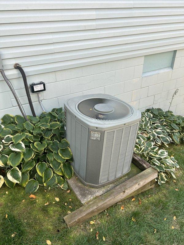 Assessed 23-year-old A/C condenser unit positioned on deteriorating wooden platform that has partially collapsed on one corner. Unit is mounted on a hillside against concrete wall with significant rot in the support structure. Recommended complete system replacement rather than attempting to level existing platform, as moving the aged unit for repairs would not be cost-effective.
