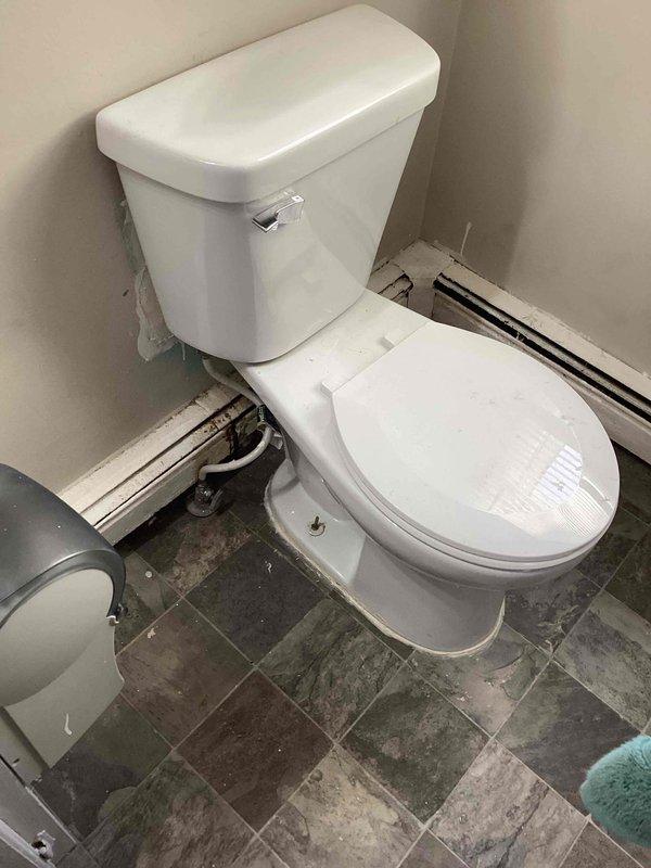 Responded to service call for toilet leaking from base and tank. Inspected two-piece white porcelain toilet positioned in bathroom corner with gray stone-look tile flooring. Presented client with options for repair or complete replacement of the malfunctioning fixture.