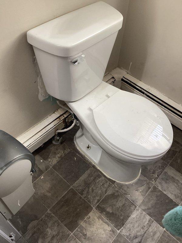Responded to service call for toilet leaking from base and tank. Inspected two-piece white porcelain toilet positioned in bathroom corner with gray stone-look tile flooring. Presented client with options for repair or complete replacement of the malfunctioning fixture.