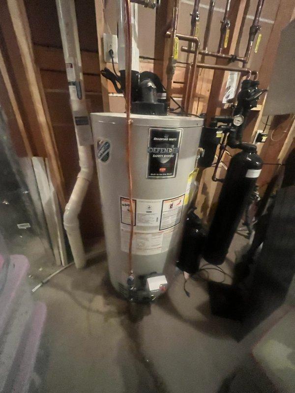 Conducted water heater inspection following customer's report of small water accumulation around unit. Assessed existing water heater with visible "Defender Safety System" and adjacent water treatment system in unfinished utility area. Determined unit requires replacement due to active leak; recommended installation of new water heater with expansion tank under Family Rewards program.