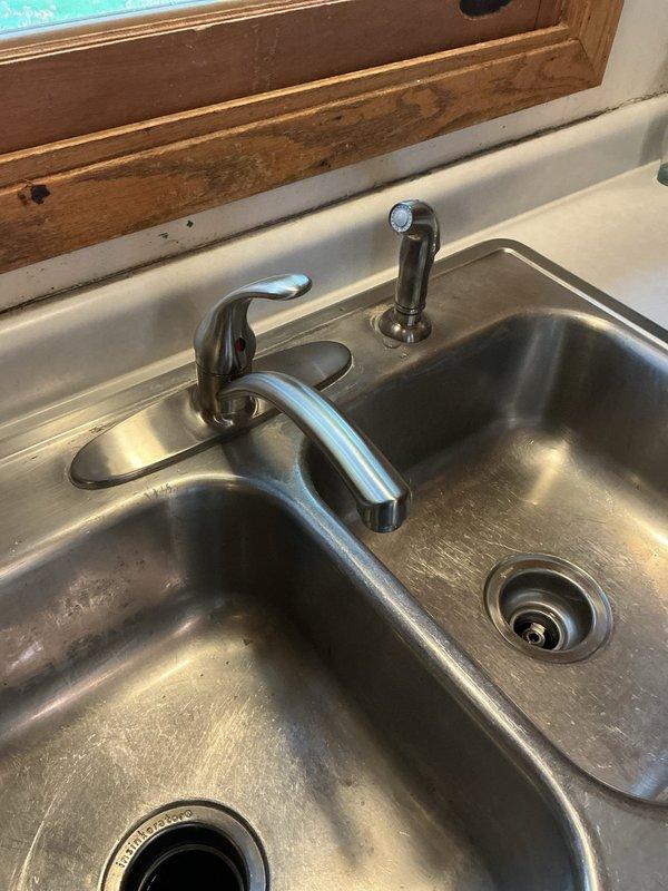Successfully replaced customer-supplied kitchen faucet after the previous unit had become stiff and difficult to operate. Installed new chrome-finished faucet with single lever handle and separate spray nozzle attachment on stainless steel double basin sink, ensuring proper functionality and leak-free operation.