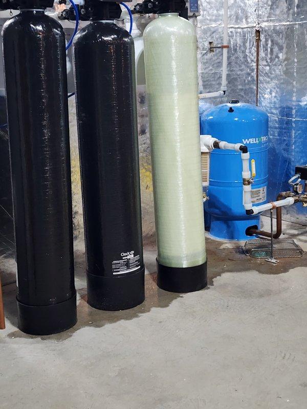 Responded to service call regarding water leakage around water treatment system comprising three vertical tanks (two black filtration units and one green nitrate filter). Upon inspection, identified a crack in the drain adapter on the nitrate filter as the source of the leak. Replaced the damaged adapter with a new component from inventory, resolving the water leakage issue.