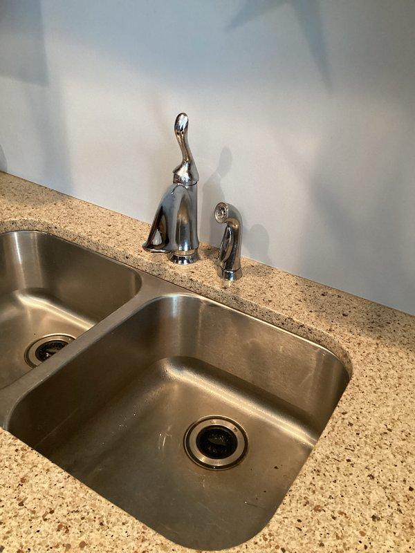 Successfully installed customer-supplied kitchen faucet in a double stainless steel sink mounted in beige granite/composite countertop. Completed installation of main chrome fixture with high arched neck and single lever handle, ensuring proper function and no leaks.