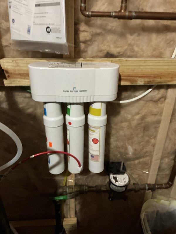Completed annual plumbing inspection revealing two non-functioning fixtures: kitchen sink spigot and water line to refrigerator. Documented existing water heater installation with copper and PVC connections in utility area, and inspected wall-mounted Water Factory Systems filtration unit with three filter cartridges (blue, green, and yellow tops) that appears to be in working condition.
