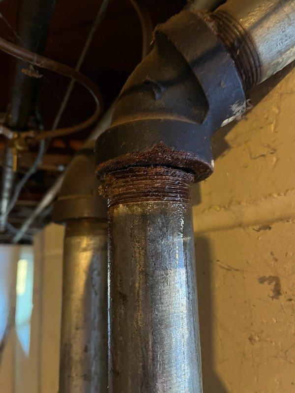 Attended property to diagnose basement leak located at galvanized pipe junction below kitchen sink where it connects to main sewer line. Visual inspection confirmed significant corrosion and rust at threaded connections of cast iron elbow fitting joining vertical and horizontal pipes. Conducted assessment and sold replacement estimate as existing galvanized piping has rusted through, requiring complete replacement. Scheduled return visit to perform full pipe replacement.