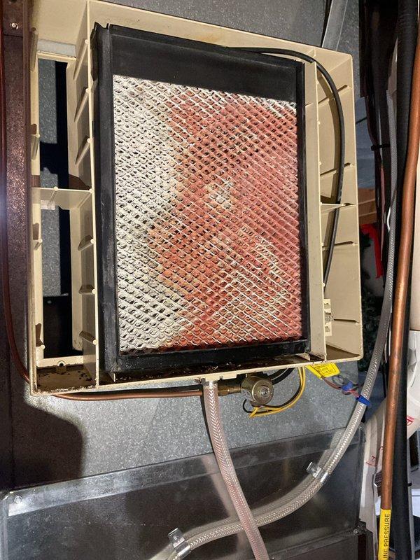 Responded to service call regarding water accumulation around furnace installed in October. Inspection revealed humidifier completely filled with mineral deposits causing water overflow. Also identified improper installation where AC drain line clamp screw had penetrated through flue pipe creating potential safety hazard. Replaced humidifier pad and recommended proper rerouting of humidifier water line to hot water supply with removal of piercing valve.