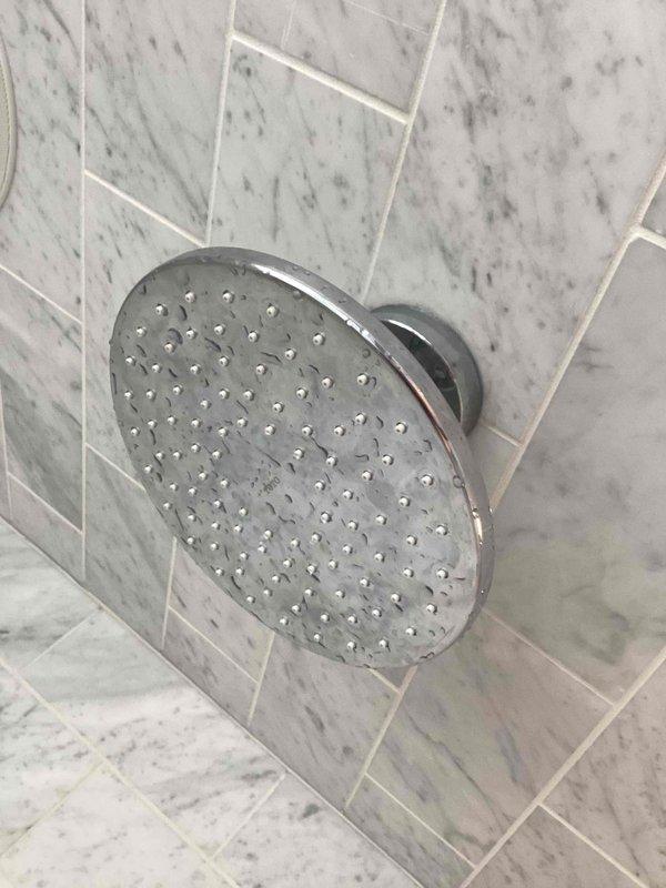 Diagnosed low water pressure in upstairs shower caused by significant mineral buildup and corrosion inside the shower head. Inspected the premium rainfall-style chrome fixture mounted on marble tile wall, noting extensive calcium deposits blocking multiple water outlet holes. Provided client with options for descaling or replacement to restore proper water flow.