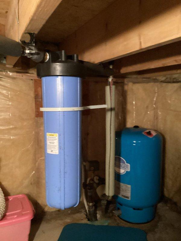 Inspected whole house water filtration system located in crawl space. Diagnosed issues with cold water temperature and evaluated sediment filter that customer was unable to remove. Documented current setup including blue cylindrical filter housing mounted to wooden joists and connected to pressure tank system for future filter replacement recommendation.