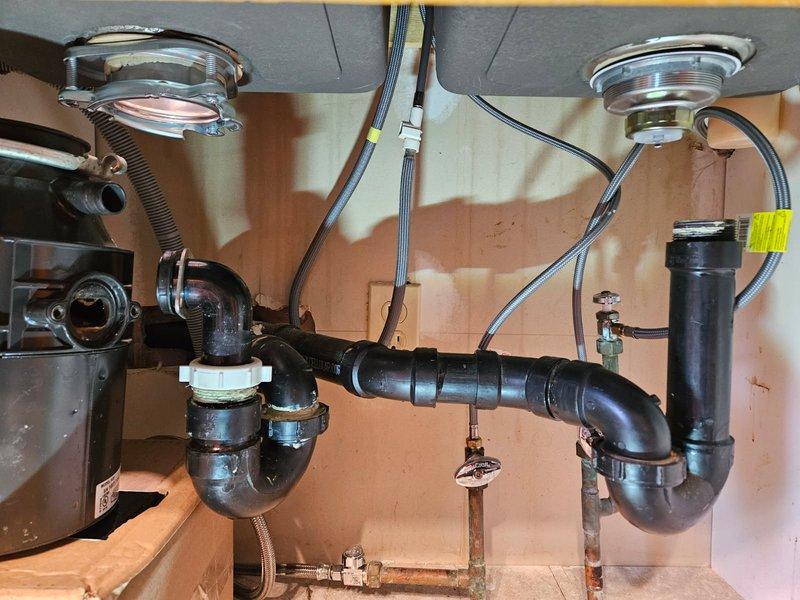 Completed kitchen sink drain installation by cutting and realigning kitchen drain pipe to properly connect with new countertop configuration. Successfully installed and connected dual-basin drain assembly with P-trap configuration, ensuring proper alignment and leak-free operation for both sink drains and disposal hookup.