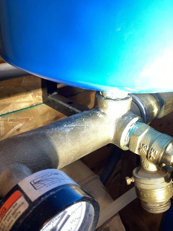 Responded to water leak in mechanical room originating from well system's pressure control. Upon arrival, found variable speed pump manifold cracked and leaking, with pump already shut off to prevent further water discharge. Documented existing plumbing configuration showing blue pressure tank, brass piping with visible moisture accumulation, and gray pump control unit with connected PVC and shutoff valves. Provided estimate for manifold replacement to restore system functionality.