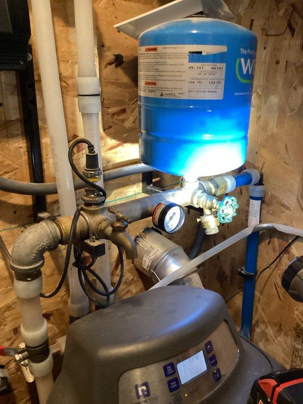 Responded to water leak in mechanical room originating from well system's pressure control. Upon arrival, found variable speed pump manifold cracked and leaking, with pump already shut off to prevent further water discharge. Documented existing plumbing configuration showing blue pressure tank, brass piping with visible moisture accumulation, and gray pump control unit with connected PVC and shutoff valves. Provided estimate for manifold replacement to restore system functionality.
