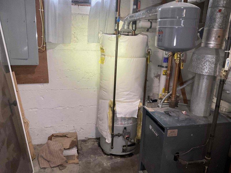 Responded to service call for reported water heater leak in basement utility area. Upon inspection, identified a 1998 model 50-gallon natural gas water heater actively leaking from the bottom of the tank. Conducted thorough assessment of the unit and determined the leak was not repairable due to tank failure and age of the equipment. Documented the existing installation configuration including associated plumbing connections, expansion tank, and heating system components through photographic documentation. Provided replacement estimates to homeowner for new water heater installation.
