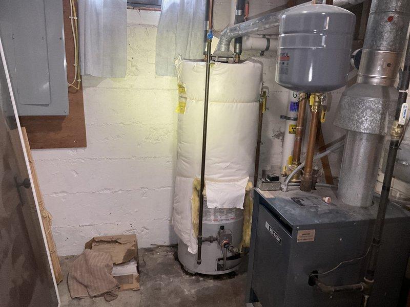 Responded to service call for reported water heater leak in basement utility area. Upon inspection, identified a 1998 model 50-gallon natural gas water heater actively leaking from the bottom of the tank. Conducted thorough assessment of the unit and determined the leak was not repairable due to tank failure and age of the equipment. Documented the existing installation configuration including associated plumbing connections, expansion tank, and heating system components through photographic documentation. Provided replacement estimates to homeowner for new water heater installation.