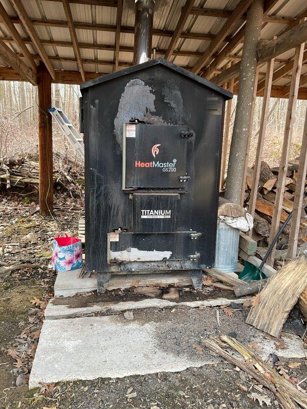 Performed comprehensive evaluation of residential heating system for new homeowner unfamiliar with boiler operation. Inspected HeatMaster GS200 Titanium outdoor wood-burning furnace located in open-sided shelter structure. Unit displayed significant paint deterioration and surface wear on upper sections, exposing underlying metal substrate. Discovered critical freeze damage to outdoor furnace cabinet, with housing compromised and broken open due to freezing conditions. Assessed indoor mechanical room containing hydronic heating distribution system with multi-zone manifold assembly, including multiple zone valves with blue handles, pressure gauges, and copper piping network. Evaluated green circulation pump control system and associated plumbing connections. Documented existing system configuration including reflective insulation installation, PVC and copper piping arrangements, and manifold pressure monitoring equipment. Coordinated with sales department to schedule estimate for replac