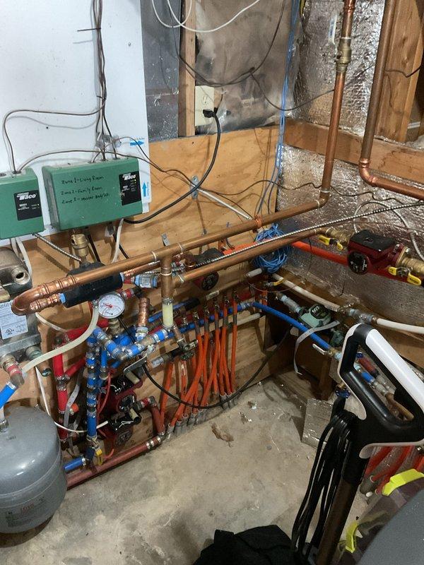 Performed comprehensive evaluation of residential heating system for new homeowner unfamiliar with boiler operation. Inspected HeatMaster GS200 Titanium outdoor wood-burning furnace located in open-sided shelter structure. Unit displayed significant paint deterioration and surface wear on upper sections, exposing underlying metal substrate. Discovered critical freeze damage to outdoor furnace cabinet, with housing compromised and broken open due to freezing conditions. Assessed indoor mechanical room containing hydronic heating distribution system with multi-zone manifold assembly, including multiple zone valves with blue handles, pressure gauges, and copper piping network. Evaluated green circulation pump control system and associated plumbing connections. Documented existing system configuration including reflective insulation installation, PVC and copper piping arrangements, and manifold pressure monitoring equipment. Coordinated with sales department to schedule estimate for replac