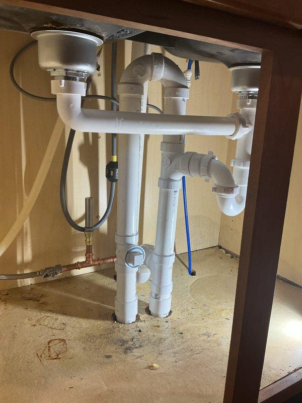 Reinstalled complete plumbing assembly underneath double-basin kitchen sink following previous removal. Reconnected water supply lines and drain system, including P-trap configuration, white PVC drain lines, and garbage disposal unit with associated discharge piping. Verified all connections secure, tested system for proper drainage and confirmed no leaks present.