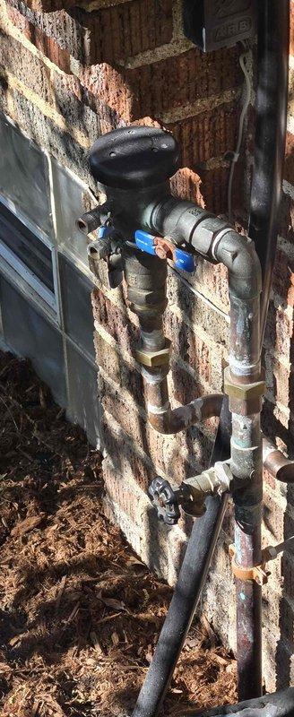 Completed plumbing inspection to diagnose reduced water pressure in sprinkler system following recent boiler installation. Inspected exterior irrigation plumbing assembly including backflow preventer, ball valves, and associated piping mounted on exterior brick wall. Documented condition of outdoor plumbing components showing weathering and age-related deterioration, and assessed system configuration to identify potential pressure loss points related to recent home modifications.