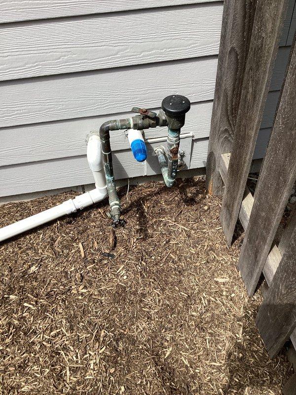 Responded to irrigation system leak involving broken piping in both exterior and interior locations. Inspected outdoor plumbing assembly featuring white PVC connected to multiple metal valves and fittings with blue-handled control valve, mounted against building exterior at grade level. Traced system through basement utility area where vertical white PVC pipe connects to horizontal copper supply lines at ceiling joists. Identified compromised pipe sections in the unusual configuration requiring replacement. Documented damage and provided part order form for estimate to repair broken pipes in both locations.
