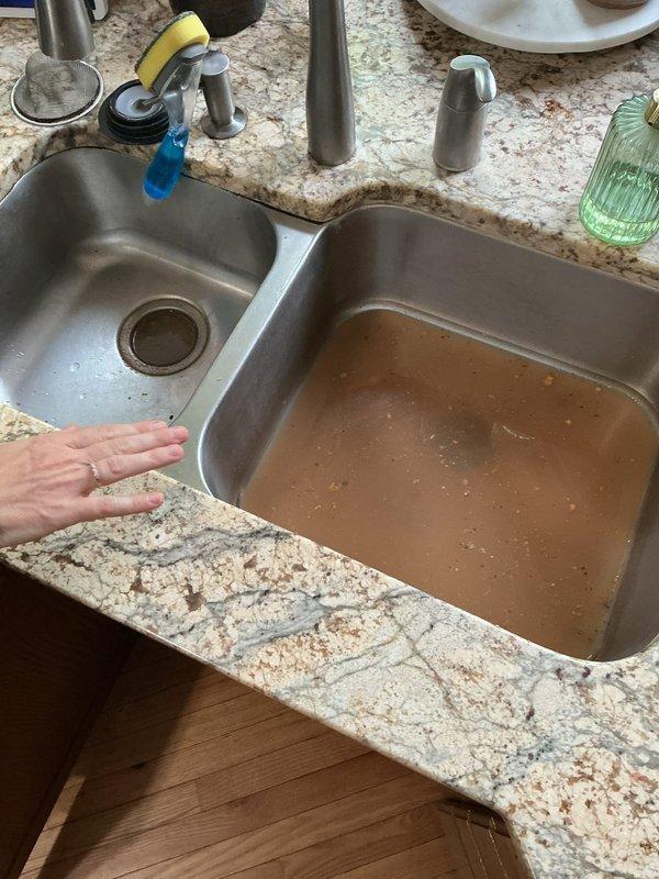 Responded to a clogged kitchen sink with complete drainage blockage on the disposal side. Upon arrival, found standing water with brown discoloration and sediment in the right basin of a double-basin stainless steel sink. Diagnosed blockage as grease accumulation in the drain line. Set up drain cable machine beneath the sink and successfully cabled the line to clear the obstruction, restoring full drainage function to the disposal side of the sink.