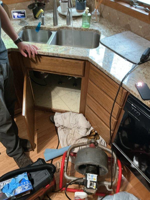 Responded to a clogged kitchen sink with complete drainage blockage on the disposal side. Upon arrival, found standing water with brown discoloration and sediment in the right basin of a double-basin stainless steel sink. Diagnosed blockage as grease accumulation in the drain line. Set up drain cable machine beneath the sink and successfully cabled the line to clear the obstruction, restoring full drainage function to the disposal side of the sink.