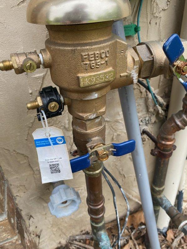 Responded to a water leak at the irrigation system connection point. Upon inspection, identified a cracked FEBCO vacuum breaker (model LK9535C) with a split body that was causing water leakage when the sprinkler system was activated. The backflow prevention device showed significant damage and was deemed non-repairable. Documented the existing installation with photographs showing the brass valve assembly, blue-handled ball valves, and copper pipe connections mounted on the exterior wall. Ordered a replacement vacuum breaker unit and will return to complete the installation once the part arrives.