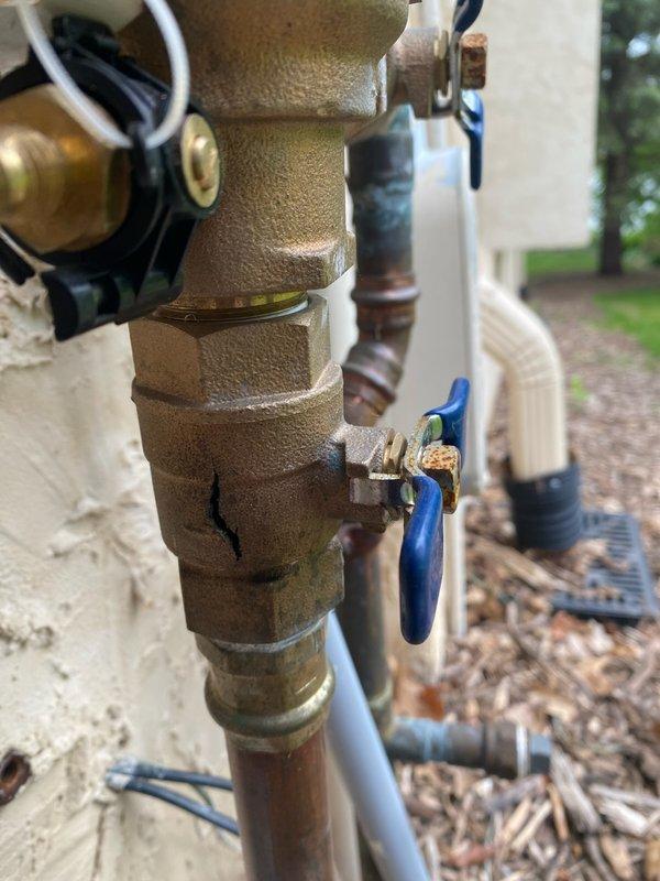 Responded to a water leak at the irrigation system connection point. Upon inspection, identified a cracked FEBCO vacuum breaker (model LK9535C) with a split body that was causing water leakage when the sprinkler system was activated. The backflow prevention device showed significant damage and was deemed non-repairable. Documented the existing installation with photographs showing the brass valve assembly, blue-handled ball valves, and copper pipe connections mounted on the exterior wall. Ordered a replacement vacuum breaker unit and will return to complete the installation once the part arrives.