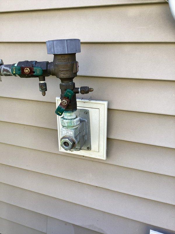 Responded to service call for leaking outdoor backflow preventer located near AC unit. Upon inspection, identified compromised Watts backflow device with active water leak. Assessment revealed missing plug component from previous winterization/blow-out service performed by irrigation company. Documented existing plumbing assembly configuration including pressure regulator, multi-valve manifold system, and backflow prevention device mounted on exterior access panel. Provided estimate for repair options and advised homeowner of requirement to obtain HOA approval prior to proceeding with any modifications to the backflow system.