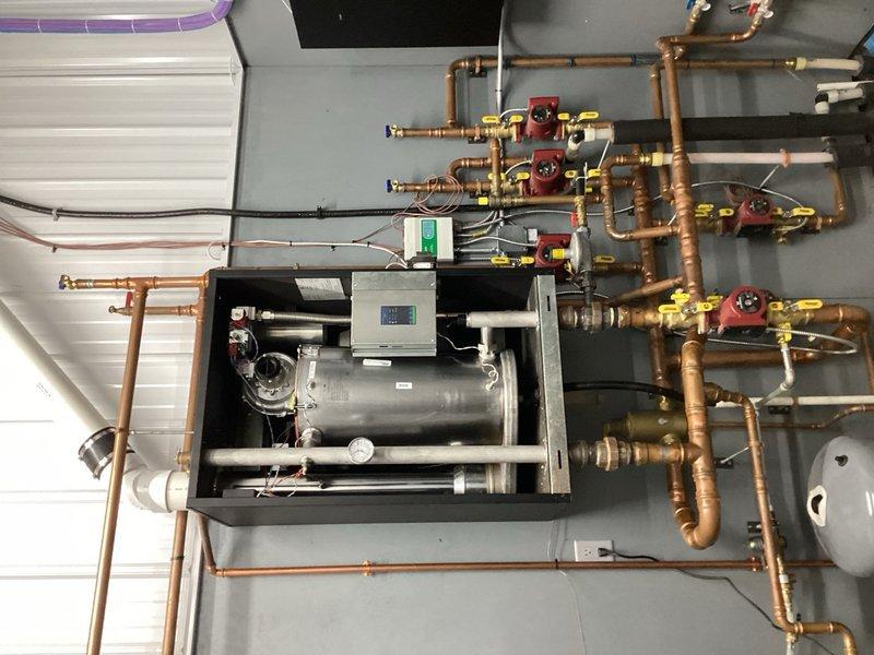 Completed preventive maintenance tune-up on HVAC equipment and hydronic heating system. Inspected boiler installation in mechanical room, documenting extensive copper piping network with multiple zone valves, expansion tank, and circulator pump assembly. Identified active leak in boiler system located in shed/shop area, evidenced by water staining on concrete floor beneath equipment. Photographed leak source and affected piping connections for estimate preparation and customer approval before proceeding with repairs.