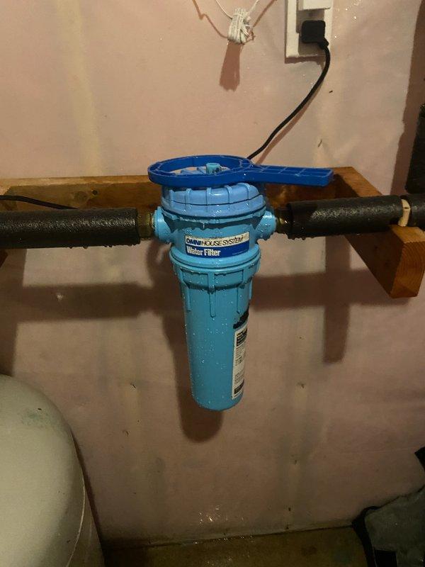 Responded to water leak emergency at residential property where home security system detected leak in water filtration system. Upon arrival, found old whole house water filter had failed and was leaking. Homeowner had already shut off main water supply to prevent further damage. Assessed the existing blue cylindrical filter unit and determined replacement was necessary. Removed failed filter assembly and installed new spin down filter with proper copper pipe connections. Mounted new white filtration unit on existing wooden backing board, secured all plumbing connections, and tested system for proper operation and leak-free performance. Restored water service to residence and verified all fixtures operating normally.