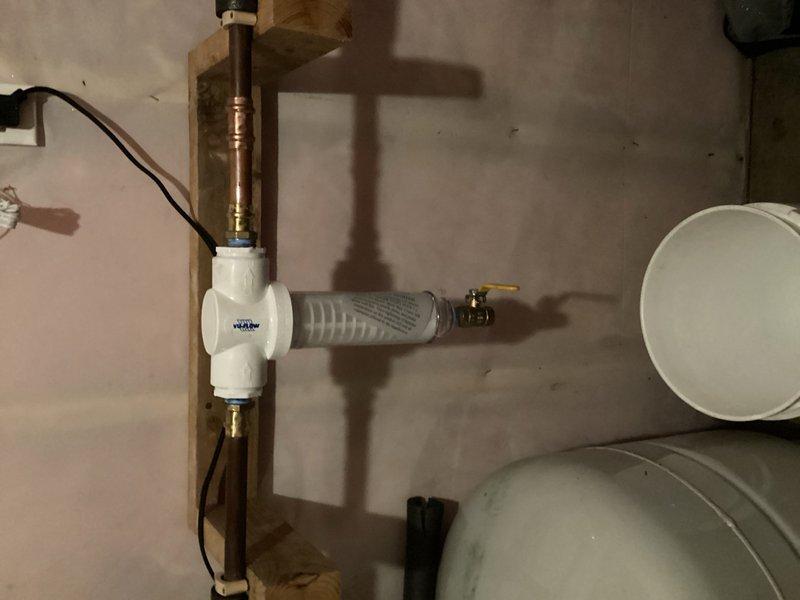 Responded to water leak emergency at residential property where home security system detected leak in water filtration system. Upon arrival, found old whole house water filter had failed and was leaking. Homeowner had already shut off main water supply to prevent further damage. Assessed the existing blue cylindrical filter unit and determined replacement was necessary. Removed failed filter assembly and installed new spin down filter with proper copper pipe connections. Mounted new white filtration unit on existing wooden backing board, secured all plumbing connections, and tested system for proper operation and leak-free performance. Restored water service to residence and verified all fixtures operating normally.